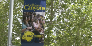 Vertical banner hanging from a light pole on a college campus. The banner features the text "College of the Canyons" in bold yellow and white letters on a blue background, along with a photo of smiling students seated in a lecture or event setting. Below the photo, the words "ALWAYS DYNAMIC" are displayed in yellow text against a stylized paintbrush-stroke background. The scene is framed by lush green tree branches in the background.